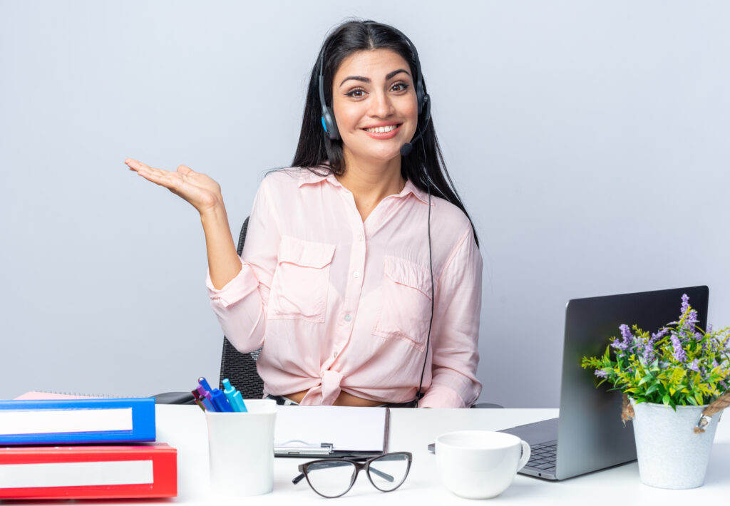 young beautiful woman in casual clothes with headphones and microphone looking at camera smiling confident presenting with arm sitting at the table with laptop over white background working in office